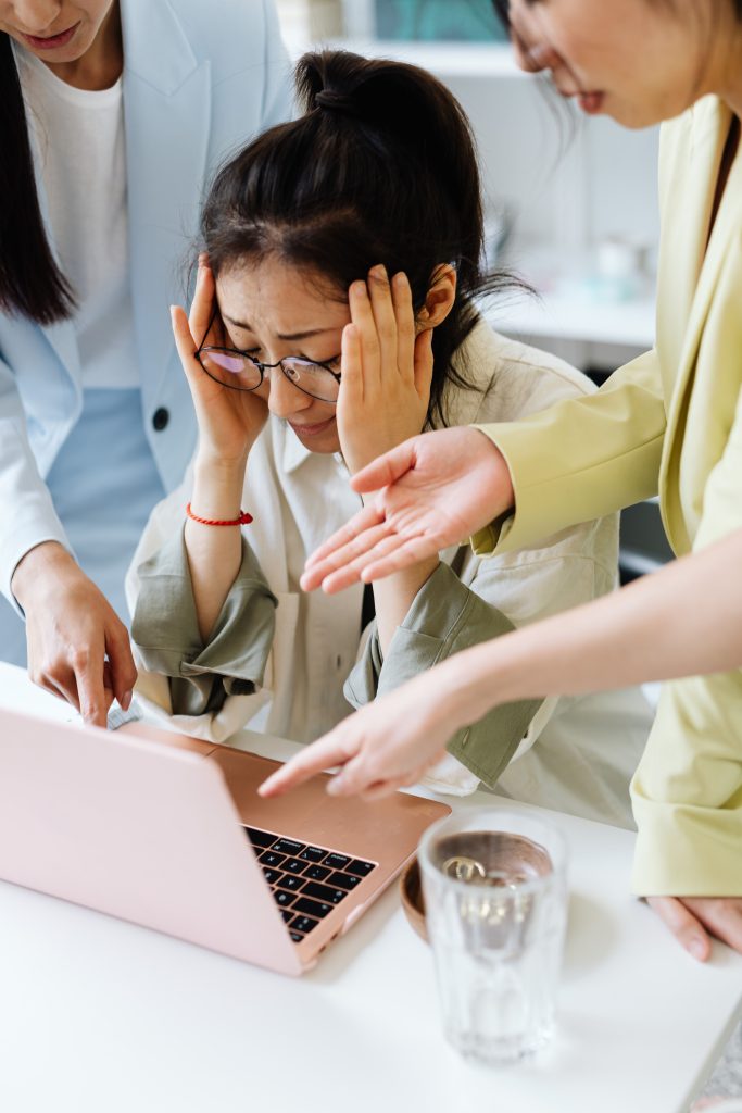 An adult woman looking stressed at work, surrounded by coworkers pointing at a laptop indoors.